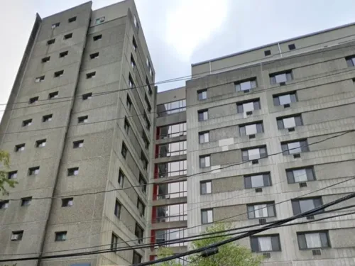 A low-angle view of Danesh Residences, a tall gray concrete brutalist building with many small windows and a central glass stairwell or connector.