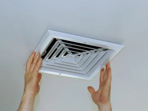 A person installing a white square ceiling air vent.