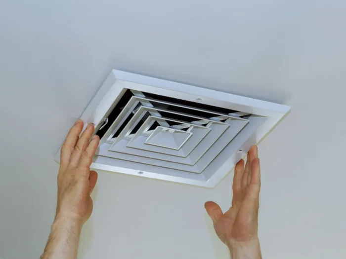 A person installing a white square ceiling air vent.