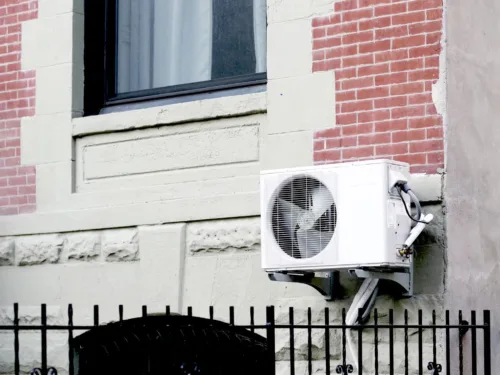 An outdoor mini-split heat pump unit installed on the side of a brick and beige stone-sided residential building.