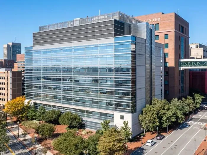 A large modern multi-story lab building featuring a reflective blue glass facade, situated in a dense urban context next to a brick building with a partial sign reading 