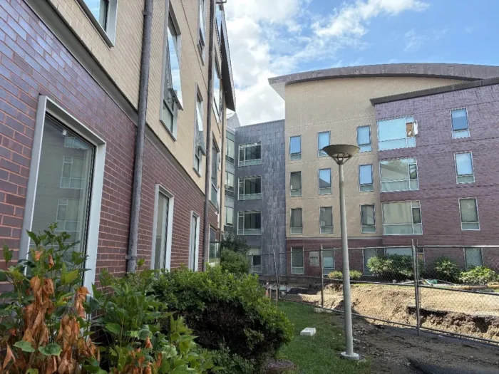 Two contemporary multi-color brick facades of West Hall surrounding a small outdoor space with a light pole and construction fencing.