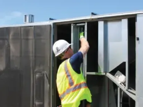 A technician in a hard hat and safety vest inspecting a large rooftop unit.