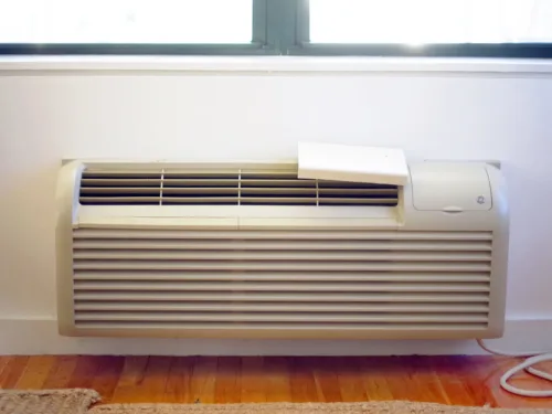 A wall-mounted PTAC heating and cooling unit installed under a window on a white wall.
