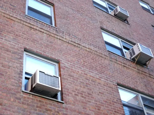 a red brick building with several windows, three of which have air conditioning units installed.