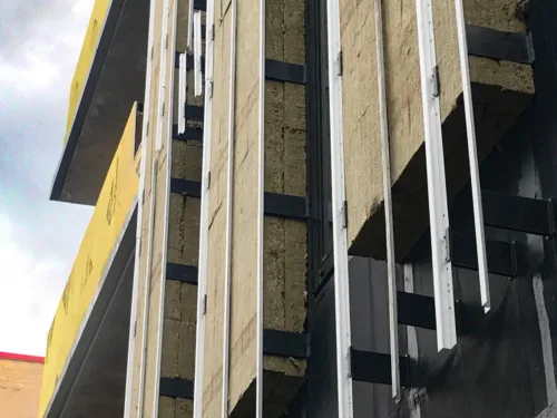 Exterior wall construction featuring thick, fibrous insulation batts secured between vertical metal framing rails, with yellow sheathing visible on the adjacent wall.