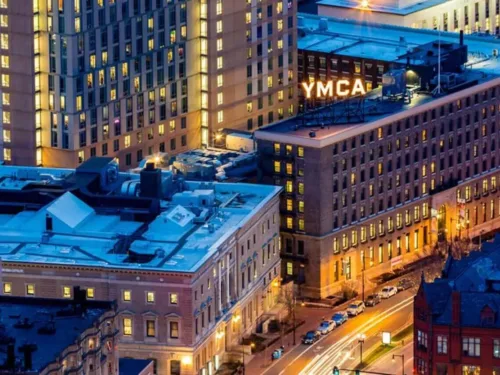 An urban scene at dusk showing a multi-story brick YMCA building with its rooftop sign illuminated in yellow.