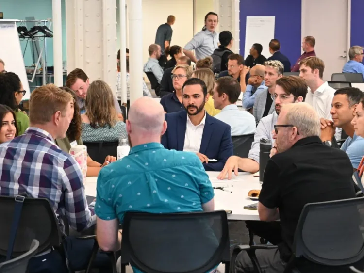 A diverse group of people gathered around a long white table in a modern, brightly lit meeting room, engaged in a discussion.